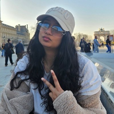 Sana in a cap and sunglasses, throwing a peace sign outside the Louvre